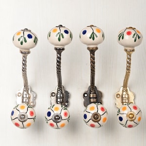 May include: Four wall hooks featuring cream-colored ceramic knobs with hand-painted green leaves and multi-colored dots. Each hook has a twisted metal stem in varying metallic finishes, including silver, dark bronze, and gold, with a floral base.