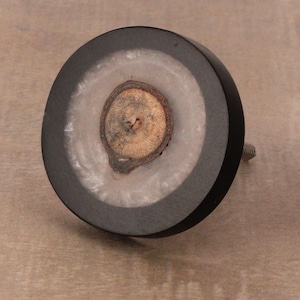May include: A round wooden knob with a black outer ring and a white center. The center features a natural wood slice with a brown and tan color.