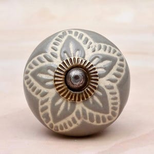 Può includere: Un pomello rotondo in ceramica per mobili di colore grigio tenue con un motivo floreale in rilievo color crema. Il centro del pomello presenta una piastra circolare in metallo color oro con una piccola sporgenza metallica scura e arrotondata.