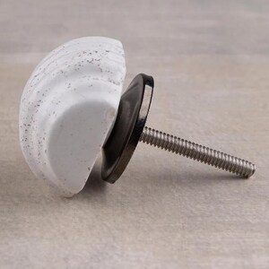 Könnte beinhalten: Weißer Keramik-Schrankknauf mit Muscheldesign. Der Knauf hat einen schwarzen Metallsockel und eine silberne Gewindeschraube. Das Muscheldesign hat ein gesprenkeltes Muster. Diese dekorative Hardware ist für Schubladen und Schränke geeignet.