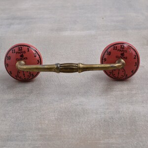 以下が含まれることがあります： 真鍮仕上げとピンクのセラミック製の顔を持つアンティーク調のキャビネットノブのペア。各ノブの顔には、「Paris Clocks」という文字と黒いローマ数字の時計が描かれています。