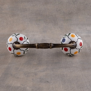 Könnte beinhalten: Ein weißer Keramik-Schubladengriff mit floralem Design mit roten, gelben und blauen Akzenten. Der Griff hat eine Messingstange, die die beiden Knöpfe verbindet.