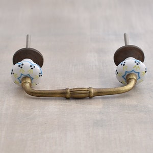 以下が含まれることがあります： 真鍮製のキャビネットプルと白いセラミックノブ。ノブには青と黄色の花柄と黒いドットがあります。