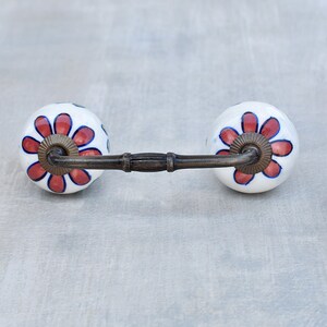 May include: White ceramic drawer pull with a red and blue floral design. The pull has a metal bar connecting the two knobs.
