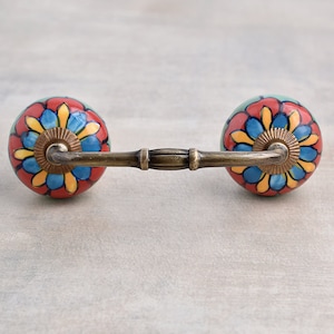 May include: A brass drawer pull with two round ceramic knobs. The knobs are decorated with a colourful floral pattern in shades of red, blue, yellow, and green.