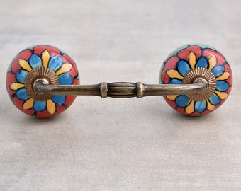 Puede incluir: Un tirador de cajón de latón con dos pomos redondos de cerámica. Los pomos están decorados con un patrón floral colorido en tonos rojos, azules, amarillos y verdes.