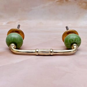 Peut inclure: Une poignée de tiroir en métal doré dotée d'une barre horizontale à la texture rappelant le bambou. La poignée est fixée à deux boutons ronds en céramique vert sauge, ornés d'un motif floral blanc et montés sur une base circulaire dorée.