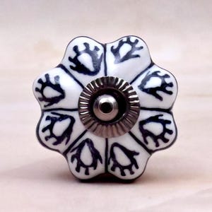 Op de afbeelding: Een witte keramische meubelknop in de vorm van een bloem met acht afgeronde bloemblaadjes. Elk bloemblaadje heeft een donkerblauw, handgetekend abstract patroon. Een cirkelvormig, geribbeld, zilverkleurig metalen onderdeel zit in het midden.