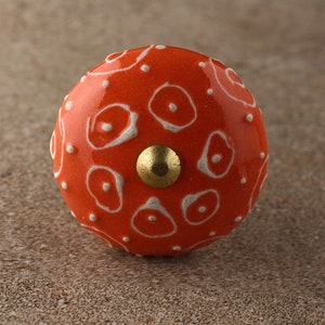 May include: Orange ceramic knob with a white abstract pattern and a gold metal center.