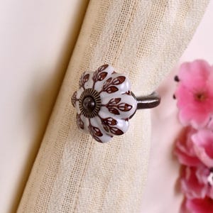 May include: A white ceramic curtain tieback featuring a scalloped flower shape with a brown leaf pattern. The center of the knob has a dark metallic circular detail, and it is attached to a curved dark metal arm that secures the beige fabric.