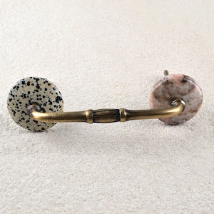 May include: A brass cabinet pull with two round, decorative knobs. One knob is white with black spots, and the other is pink with brown and white marbling.