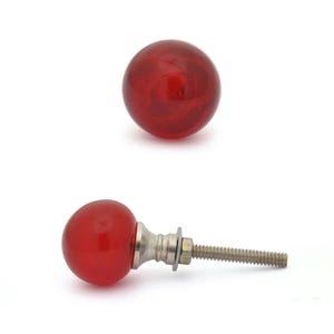May include: Two red, round cabinet knobs with a swirl pattern. One knob is shown separately, and the other is attached to a silver-toned metal base with a threaded rod, washer, and nut. The knobs are likely for furniture or decorative use.
