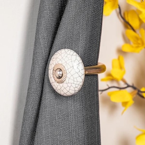 May include: White ceramic curtain tieback with a gold metal ring. The tieback has a crackled finish and a small gold button in the center.