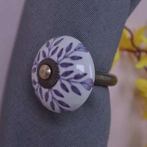 May include: A white ceramic napkin ring with a purple leaf design. The ring has a brass-colored metal base and a decorative knob. The napkin ring is holding a gray napkin. The background is blurred.