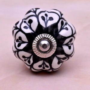 Op de afbeelding: Een ronde, gegroefde keramische meubelknop met een witte basis en een zwart handgeschilderd bloemmotief. Het midden van de knop is voorzien van een cirkelvormig zilverkleurig metalen beslag met een geribbelde rand en een gladde, bolle kern.