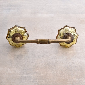 May include: A decorative cabinet pull with a brass finish and two yellow-tinted, flower-shaped glass knobs. The pull has a central bar and curved supports connecting to the knobs. The hardware is designed for furniture and adds a vintage touch.