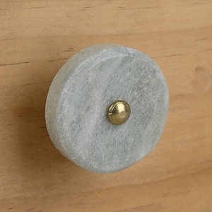 Op de afbeelding: Een ronde, lichtgrijze marmeren knop met een messing accent. De knop is gemonteerd op een lichtbruin houten oppervlak.