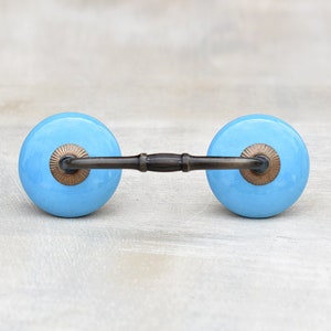 May include: A light blue ceramic drawer pull with a brass bar connecting two round knobs. The knobs have a decorative brass ring around the edge.