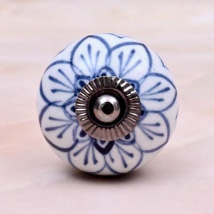 Op de afbeelding: Een ronde keramische meubelknop met een witte basis en een handgeschilderd blauw bloemenpatroon. Het midden van de knop is voorzien van een cirkelvormig, geribbeld zilverkleurig metalen onderdeel met een klein gat voor montage.