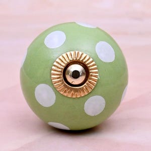 Può includere: Un pomello sferico per mobili con finitura verde chiaro caratterizzato da un motivo a pois bianchi. Il centro del pomello è accentuato da un elemento circolare in metallo dorato con un bordo scanalato e smerlato e una cupola liscia.