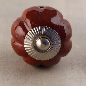 May include: A close-up of a decorative cabinet knob. The knob is a deep burgundy color with a glossy finish. It has a rounded, floral-like shape with a silver-colored metal base and center. The knob is designed for furniture.