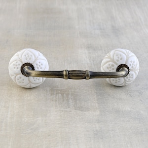May include: A white ceramic cabinet pull with a floral design on each end. The handle is a dark bronze color. The pull is centered on a light gray surface.