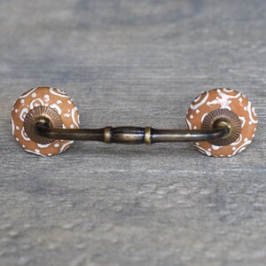 May include: A brown ceramic drawer pull with a white floral design. The pull has a metal bar connecting the two ceramic knobs.