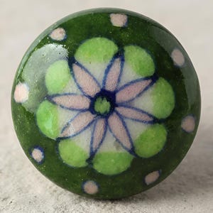 May include: A round, ceramic cabinet knob with a floral design. The knob features a dark green background with a central flower in light green and pink, outlined in blue. Small pink dots are scattered around the edge.
