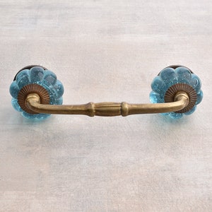 Puede incluir: Un tirador de armario de latón con dos pomos de vidrio azul. Los pomos tienen una superficie texturizada, similar a burbujas.