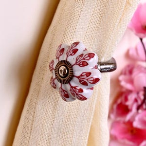 May include: A white ceramic curtain tieback featuring a scalloped flower shape with a red leaf pattern. The center of the flower has a circular bronze-toned metal accent, and the back of the tieback is attached to a dark metal hardware post.