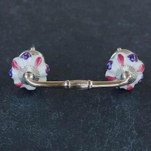 Könnte beinhalten: Weißer Keramik-Schubladengriff mit floralem Design in Rosa-, Blau- und Weißtönen. Der Griff hat eine silberne Metallstange, die die beiden Knöpfe verbindet.