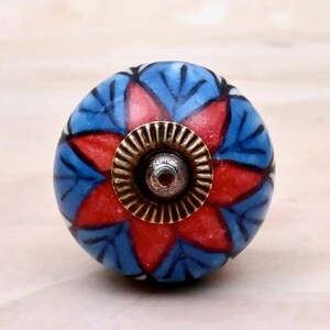 May include: A round ceramic cabinet knob featuring a vibrant floral pattern with red petals and blue leaves outlined in black. The center of the knob is adorned with a circular, ridged brass-toned metal fitting that surrounds a central silver-toned screw.