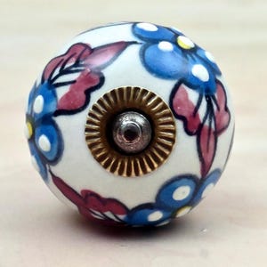 May include: A round ceramic cabinet knob with a white base featuring a hand-painted floral pattern in shades of blue and dark pink. The centre of the knob has a circular brass-toned metal plate with a ridged edge and a small silver-toned screw head.