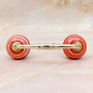 May include: A drawer pull featuring two spherical knobs with concentric orange, red, and white stripes connected by a horizontal gold-toned metal bar. The hardware includes gold-toned circular bases where the knobs meet the central connecting handle.