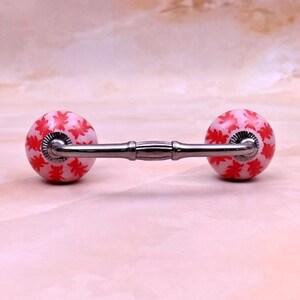 May include: A drawer pull featuring two spherical ceramic knobs with a white base and a repeating red leaf pattern. The knobs are connected by a horizontal metallic bar with a central decorative band and fluted silver-toned metal collars.
