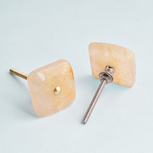 Puede incluir: Dos pomos de armario cuadrados con un acabado marmoleado marrón claro. Los pomos tienen un tornillo de metal dorado y una base de metal dorado.