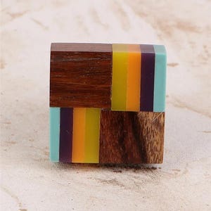 May include: A square-shaped wooden ring with colorful stripes of yellow, orange, purple, and teal. The ring is made of wood and resin, with a geometric design. The ring is a unique piece of jewelry.