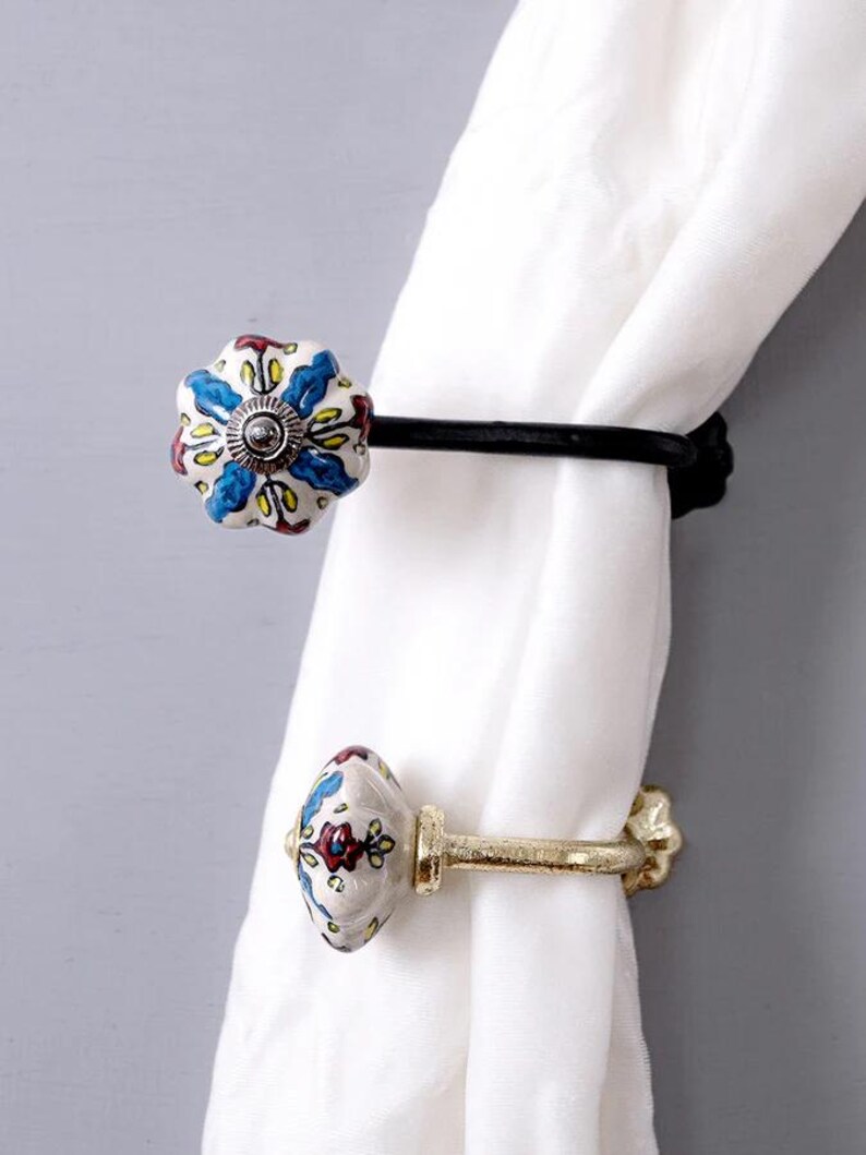 May include: Two decorative curtain tiebacks with floral patterned ceramic knobs. One tieback is black metal and the other is gold metal.