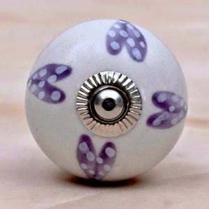 Op de afbeelding: Een ronde witte keramische meubelknop met een herhalend patroon van paarse hartvormige motieven met witte stippen. Het midden van de knop is voorzien van een gepolijste zilverkleurige metalen bevestiging met een geribbelde ronde basis.