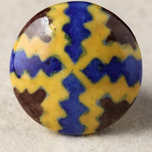 Puede incluir: Un pomo redondo de cerámica con un patrón geométrico en tonos amarillos, azules y marrones.