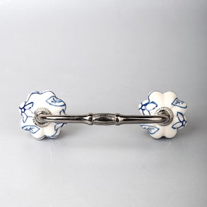 May include: A drawer pull featuring two white ceramic knobs with a blue floral pattern, connected by a polished dark metallic handle. The knobs have a scalloped, flower-like shape, and the metal bar has a central decorative ridge detail.