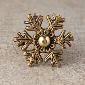 Puede incluir: Un adorno dorado en forma de copo de nieve con un centro redondo y elevado. El adorno tiene un diseño detallado con líneas y curvas intrincadas.