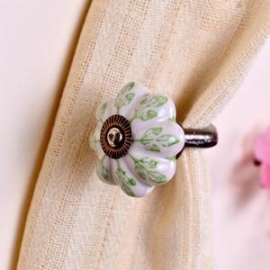 May include: A ceramic curtain tieback featuring a white, flower-shaped knob with green leaf patterns and a central bronze-toned metal accent. The tieback is secured around a piece of textured, cream-colored fabric, holding it in a gathered position.