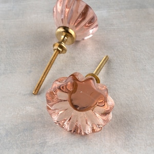 Puede incluir: Dos pomos de armario de vidrio rosa con bases de metal dorado. Los pomos tienen un diseño acanalado.