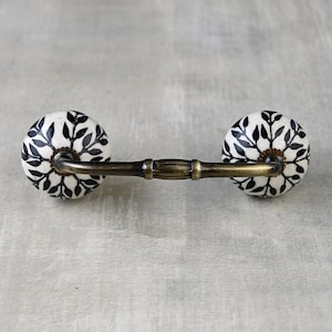 May include: A white ceramic drawer pull with a black floral design and a brass bar connecting the two knobs.