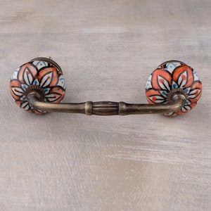 Può includere: Una maniglia per armadio decorativa con un motivo floreale. La maniglia è in metallo e ha un pomello in ceramica con sfondo bianco e un motivo floreale arancione, blu e nero.