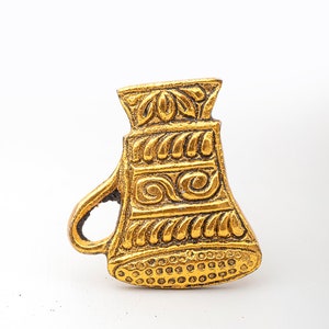 May include: A gold-colored metal pin shaped like a small pitcher or jug. The surface features embossed decorative patterns including horizontal bands of leaves, swirls, and a bottom section with a dotted texture. It has a small handle on the left side.