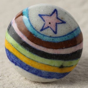 May include: A white ceramic ball with a pink star and colorful stripes in blue, yellow, green, black, brown, and pink.