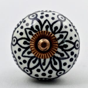 May include: A round ceramic cabinet knob featuring a white base with a black hand-painted floral pattern consisting of petals and dots. The center of the knob has a circular, ridged, metallic bronze-colored hardware component for mounting.