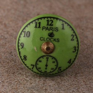May include: A green ceramic clock knob with black numbers and a smaller clock face inside. The knob says "Paris Clocks" in black lettering.